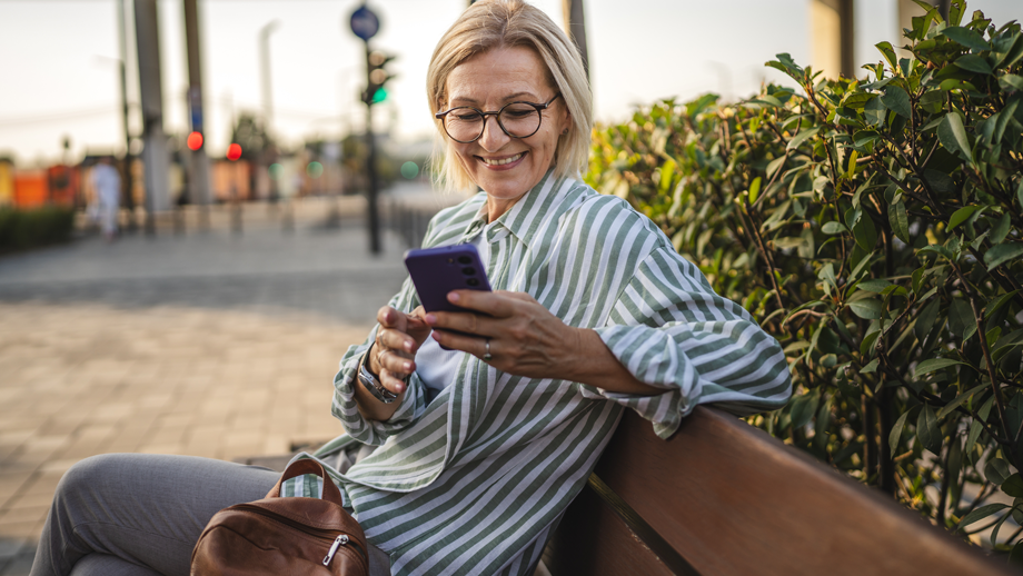 A middle-aged woman smiling while using a smartphone outdoors on a bench.