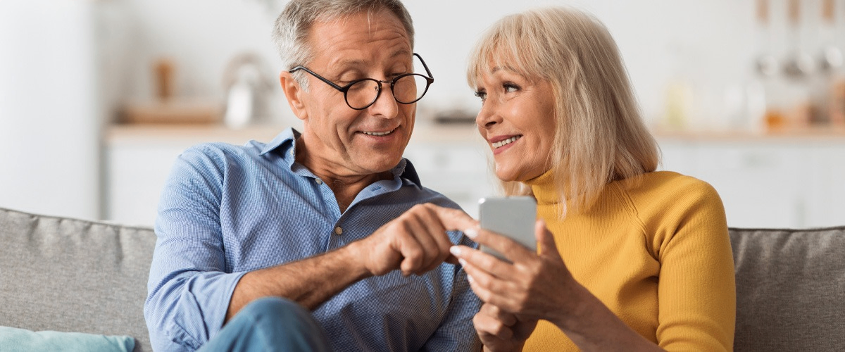 A senior couple sitting in a sofa smiling and pointing at a smart phone.
